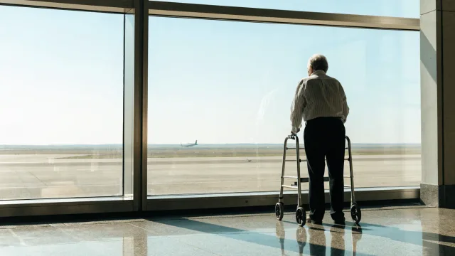 Exactly one older patient seen from behind at a runway window with a walker, standing steadily while waiting for an individual departure decision