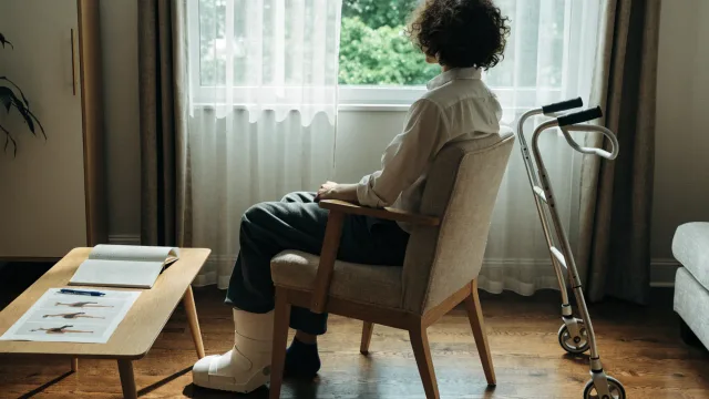 Exactly one patient seated at home for a video review, shown from behind with walking aid and exercise plan nearby, screen text absent