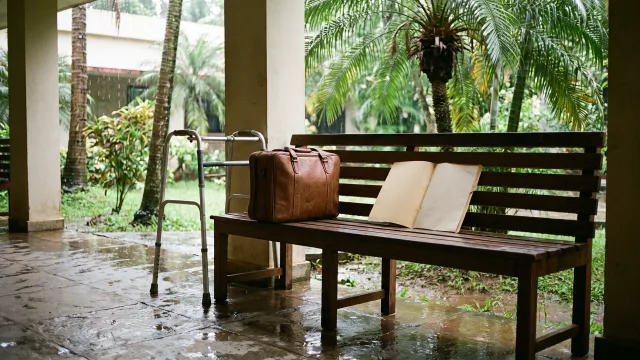 A calm Kerala hospital garden after rain with an airport carry-on, walker and joint-replacement folder on a sheltered bench, no person