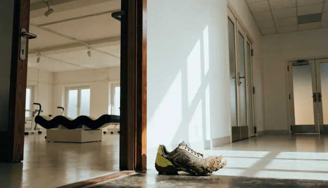 A mud-streaked football boot at the threshold between a rehabilitation gym and a surgical corridor