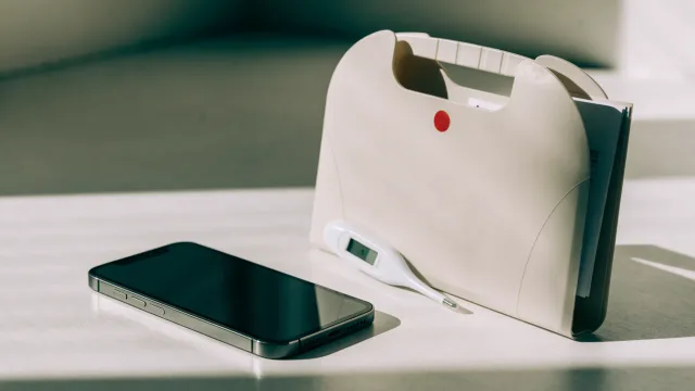 A phone beside a wound-care folder, thermometer and medicine list turned away, with one restrained red accent and no wound shown